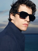 Placeholder for: Person wearing large sunglasses and a dark jacket, gazing to the side against a backdrop of a serene ocean and cloudy sky.