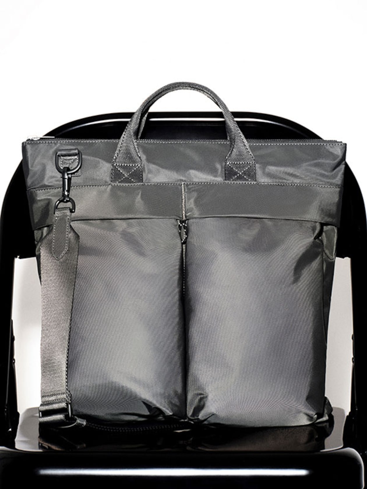 A sleek gray tote bag with dual front pockets, short handles, and a detachable shoulder strap, placed on a black chair.