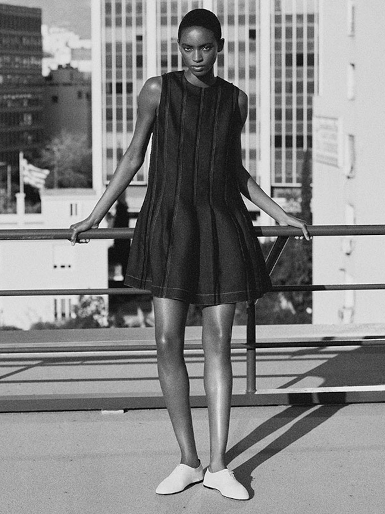 A person in a sleeveless black dress stands confidently on a rooftop, with modern buildings in the background.