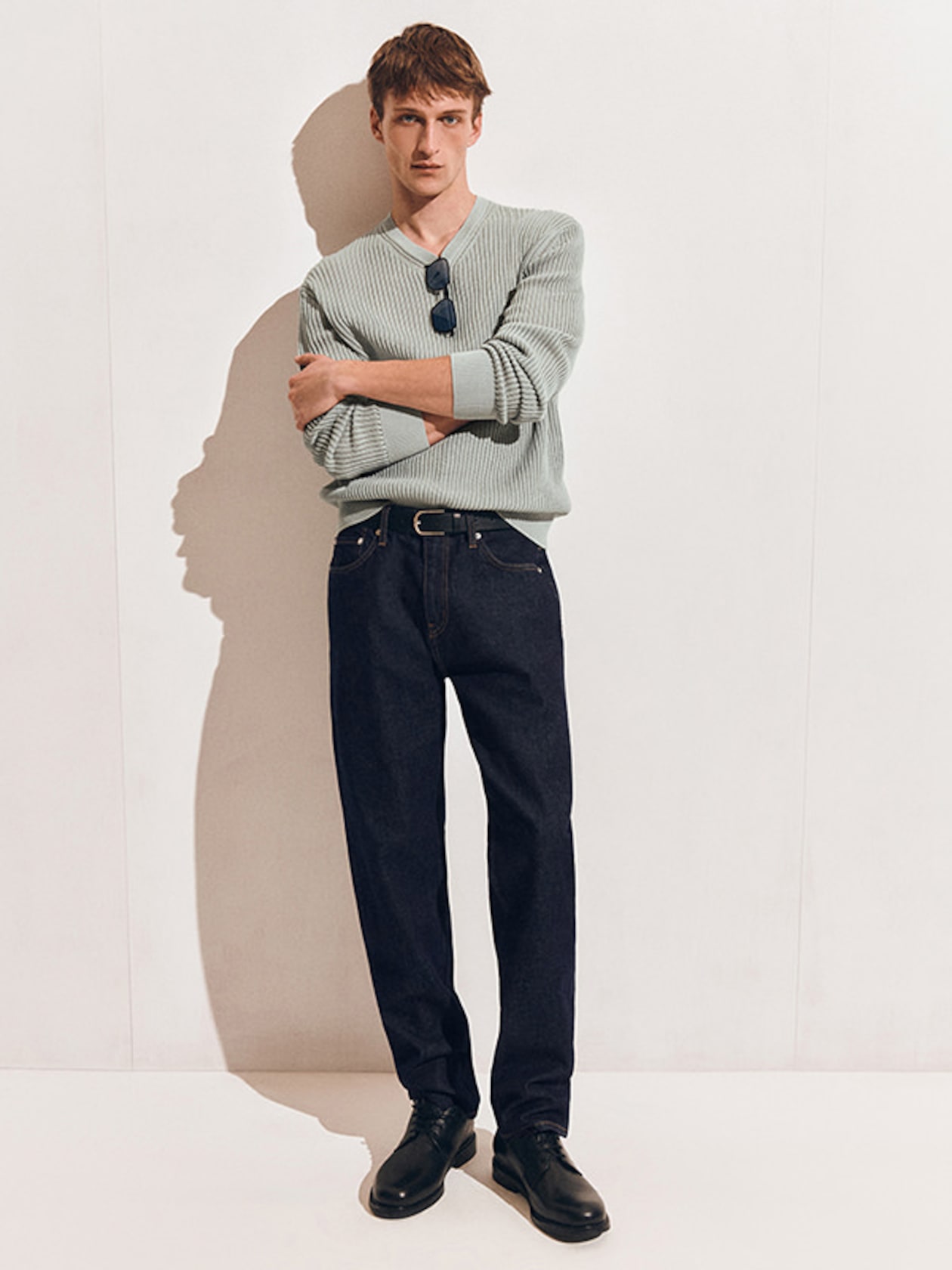 A man in a light gray sweater with sunglasses tucked in, dark jeans, and black shoes stands against a wall with arms crossed.