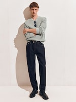 Placeholder for: A man in a light gray sweater with sunglasses tucked in, dark jeans, and black shoes stands against a wall with arms crossed.