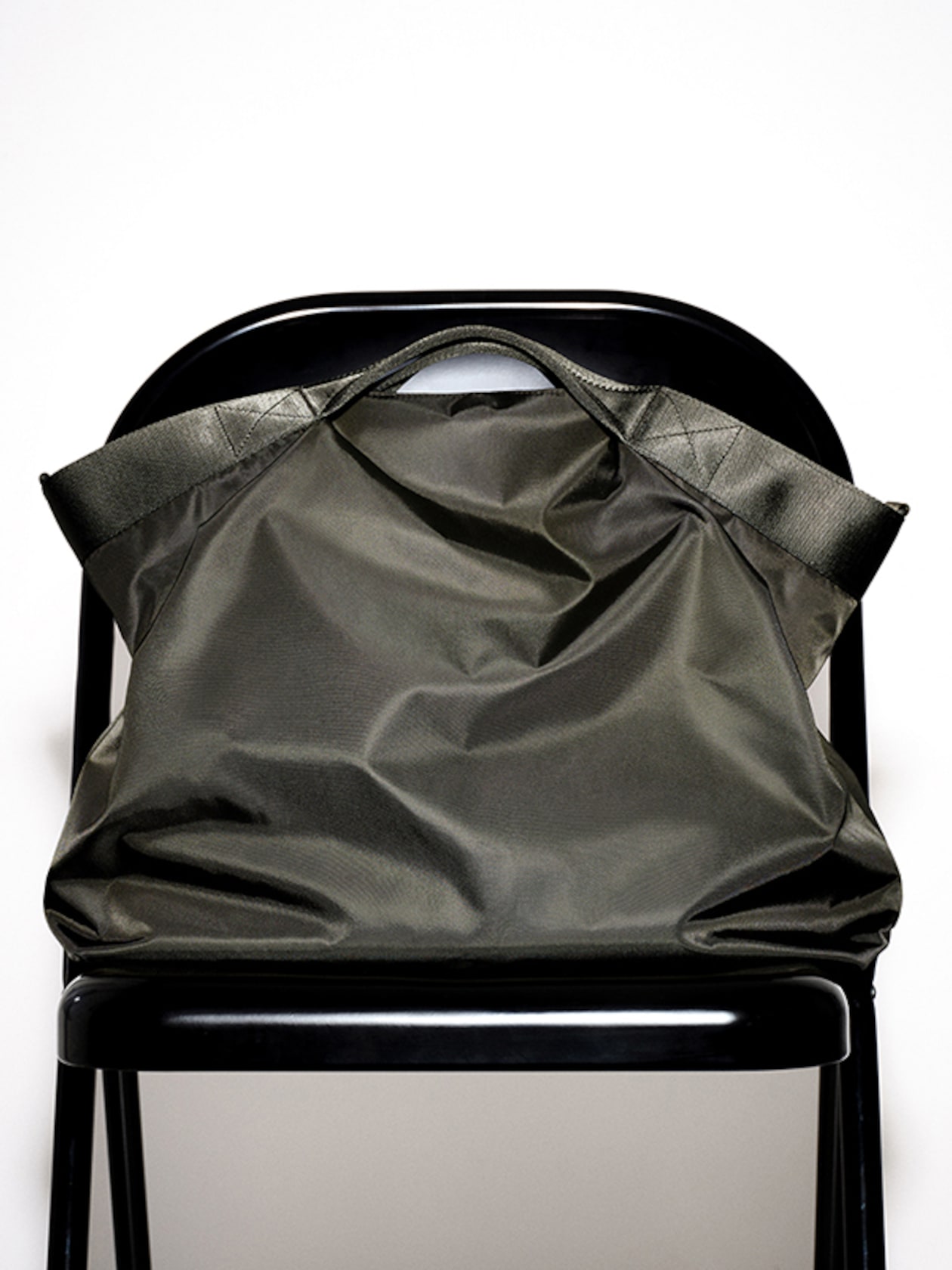 A large, dark green tote bag with black handles rests against the back of a black folding chair on a white background.