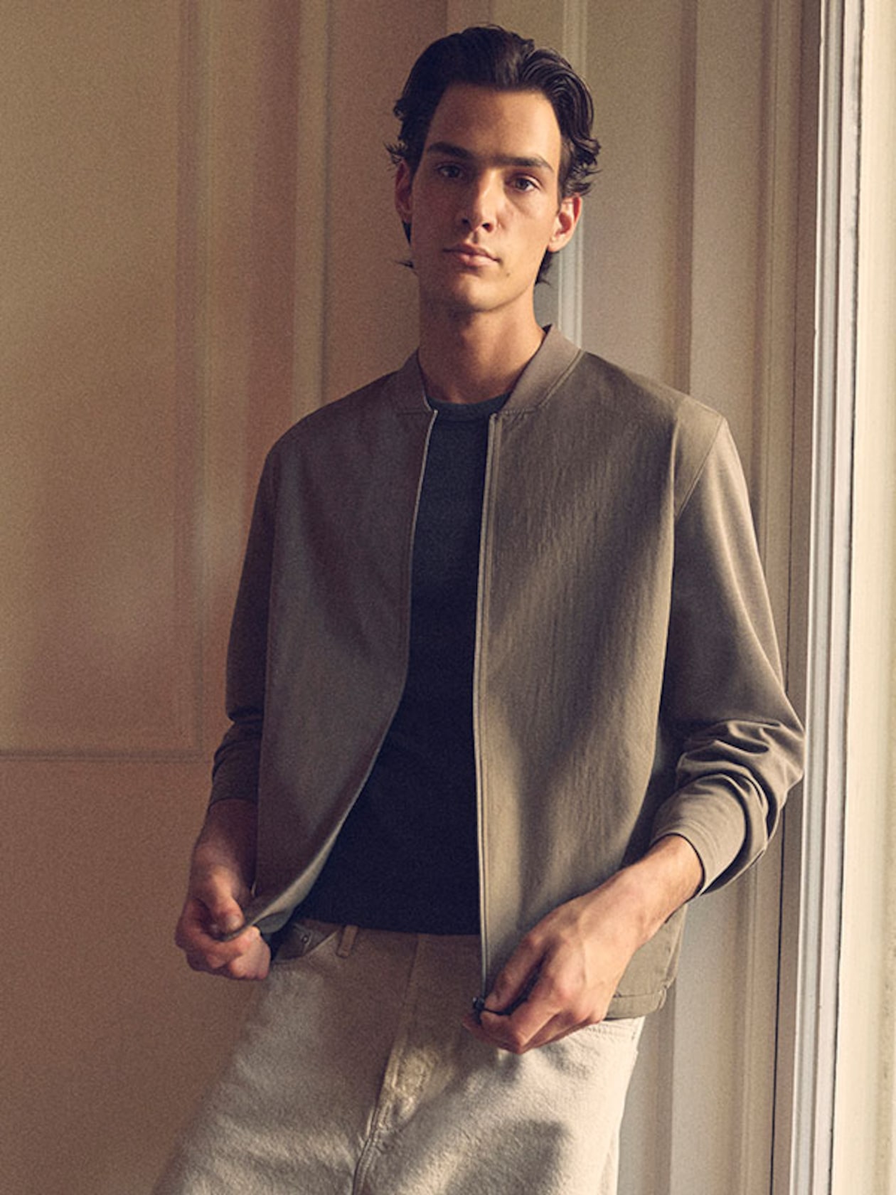 Person wearing a light jacket over a dark shirt, standing in a softly lit room with a relaxed expression.