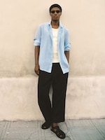 Placeholder for: Person in a light blue shirt, white tank top, black pants, and sandals, standing against a beige wall, wearing sunglasses.