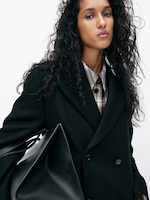 Placeholder for: Woman with wavy hair, wearing a black coat and plaid shirt, holds a large black bag against a plain white background.