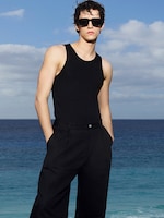 Placeholder for: Person in a black tank top and pants, wearing sunglasses, stands confidently by the ocean under a partly cloudy sky.