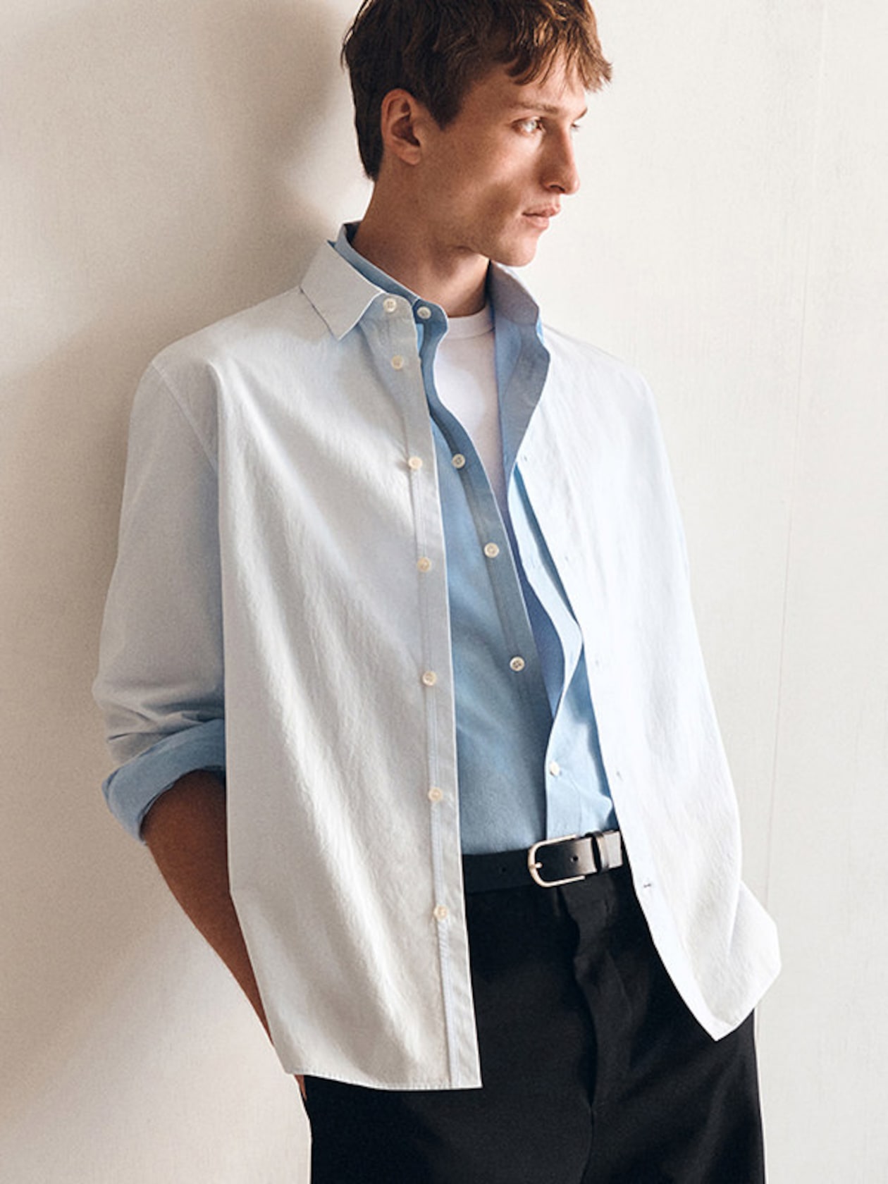 A man in a white and blue layered shirt with black pants stands against a wall, looking to the side.