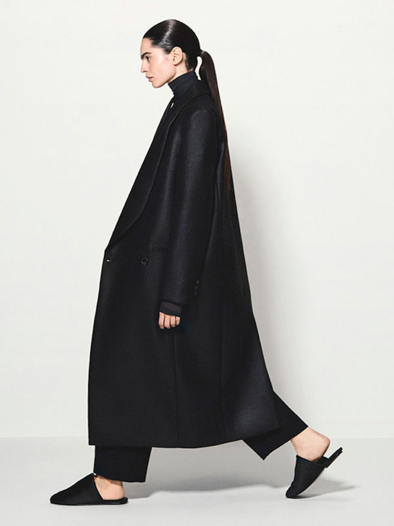 Person in profile wearing a long, black coat, black turtleneck, and black pants, walking with hair tied in a sleek ponytail.