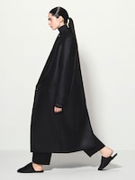 Placeholder for: Person in profile wearing a long, black coat, black turtleneck, and black pants, walking with hair tied in a sleek ponytail.
