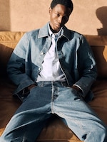 Placeholder for: Person wearing a denim jacket and jeans, sitting on a brown couch with hands in pockets, sunlight casting shadows.