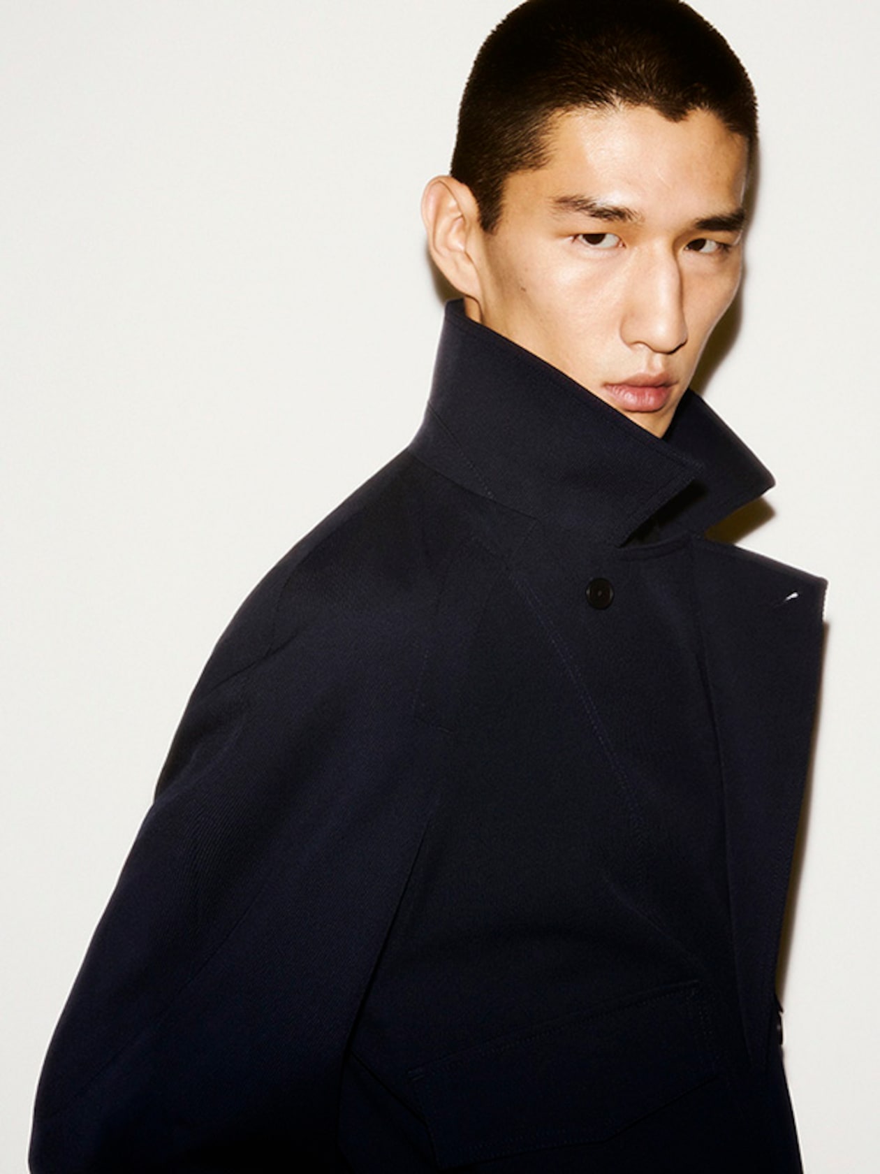 Person with short hair wearing a dark high-collared coat, looking at the camera against a light background.