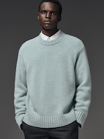 Placeholder for: Young man in a light blue sweater and white shirt, looking forward with hands in pockets, against a dark gray background.