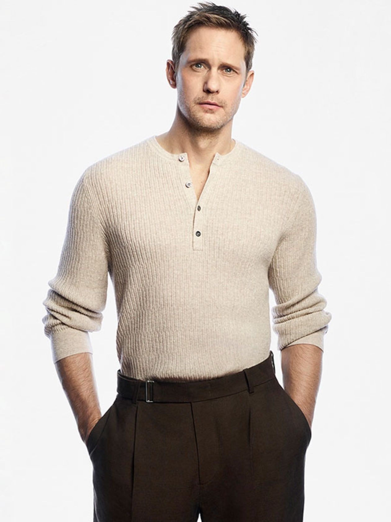 A man in a beige ribbed sweater and brown trousers stands with hands in pockets against a plain white background.
