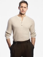 Placeholder for: A man in a beige ribbed sweater and brown trousers stands with hands in pockets against a plain white background.