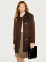 Placeholder for: Person wearing a dark coat over a pink shirt and beige skirt, holding a black textured clutch, standing against a plain background.