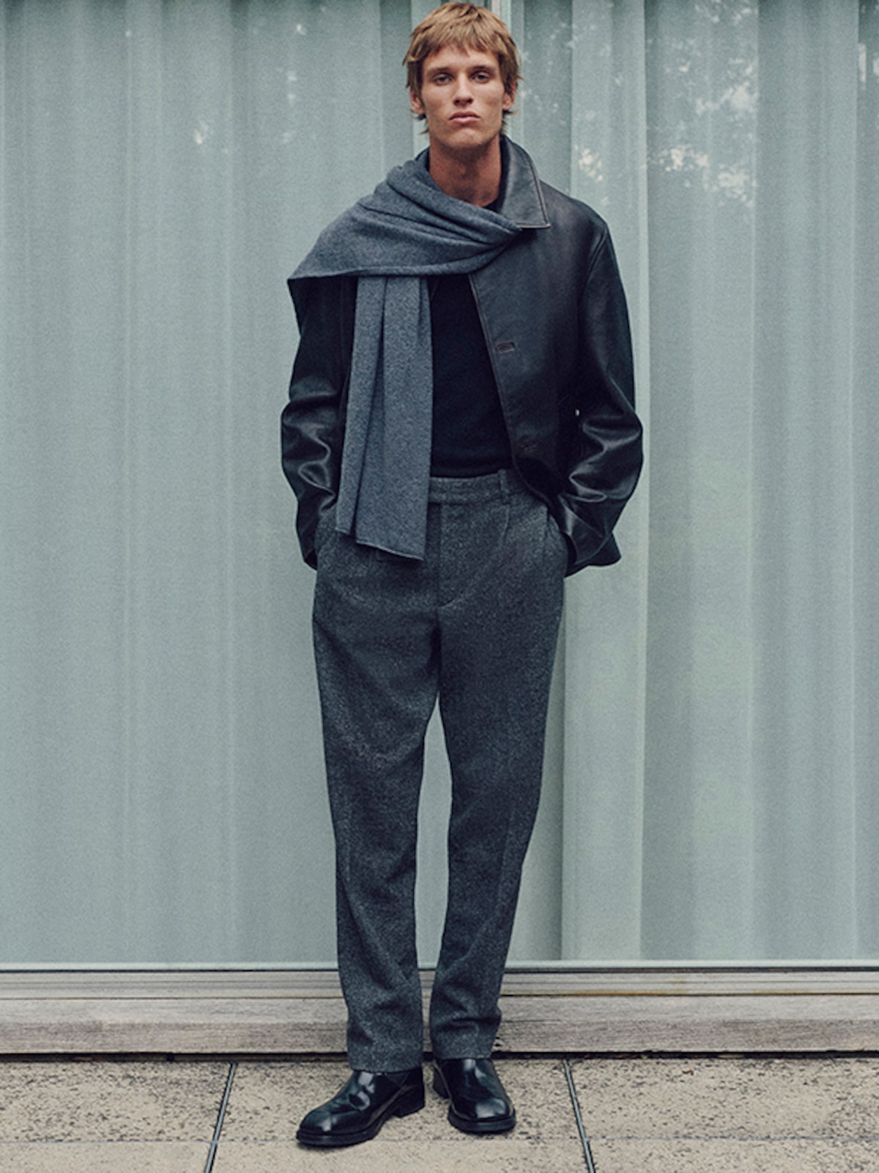 Person standing in front of a curtain, wearing a leather jacket, gray scarf, gray trousers, and black shoes, with hands in pockets.