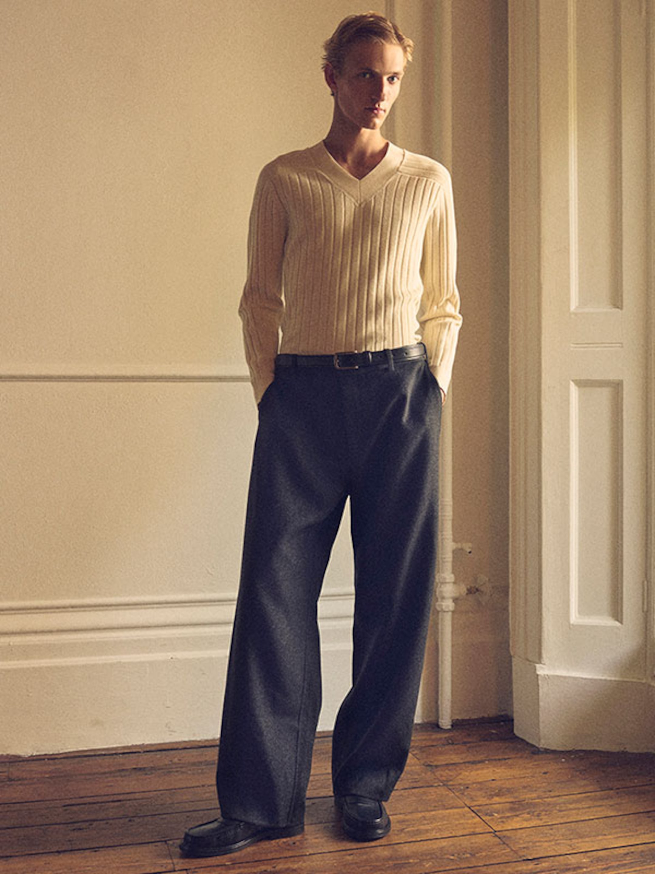 Person standing in a softly lit room wearing a cream ribbed sweater, dark wide-leg trousers, and black shoes, with a neutral expression.