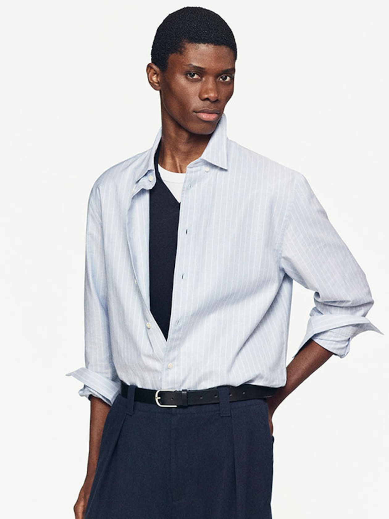 Model wearing a light blue striped shirt over a black T-shirt, with hands in pockets and a neutral expression against a plain background.