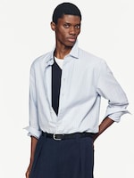 Placeholder for: Model wearing a light blue striped shirt over a black T-shirt, with hands in pockets and a neutral expression against a plain background.