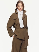 Placeholder for: Model wearing a tailored brown suit with a belted waist and large collar, complemented by a white scarf, stands against a plain background.