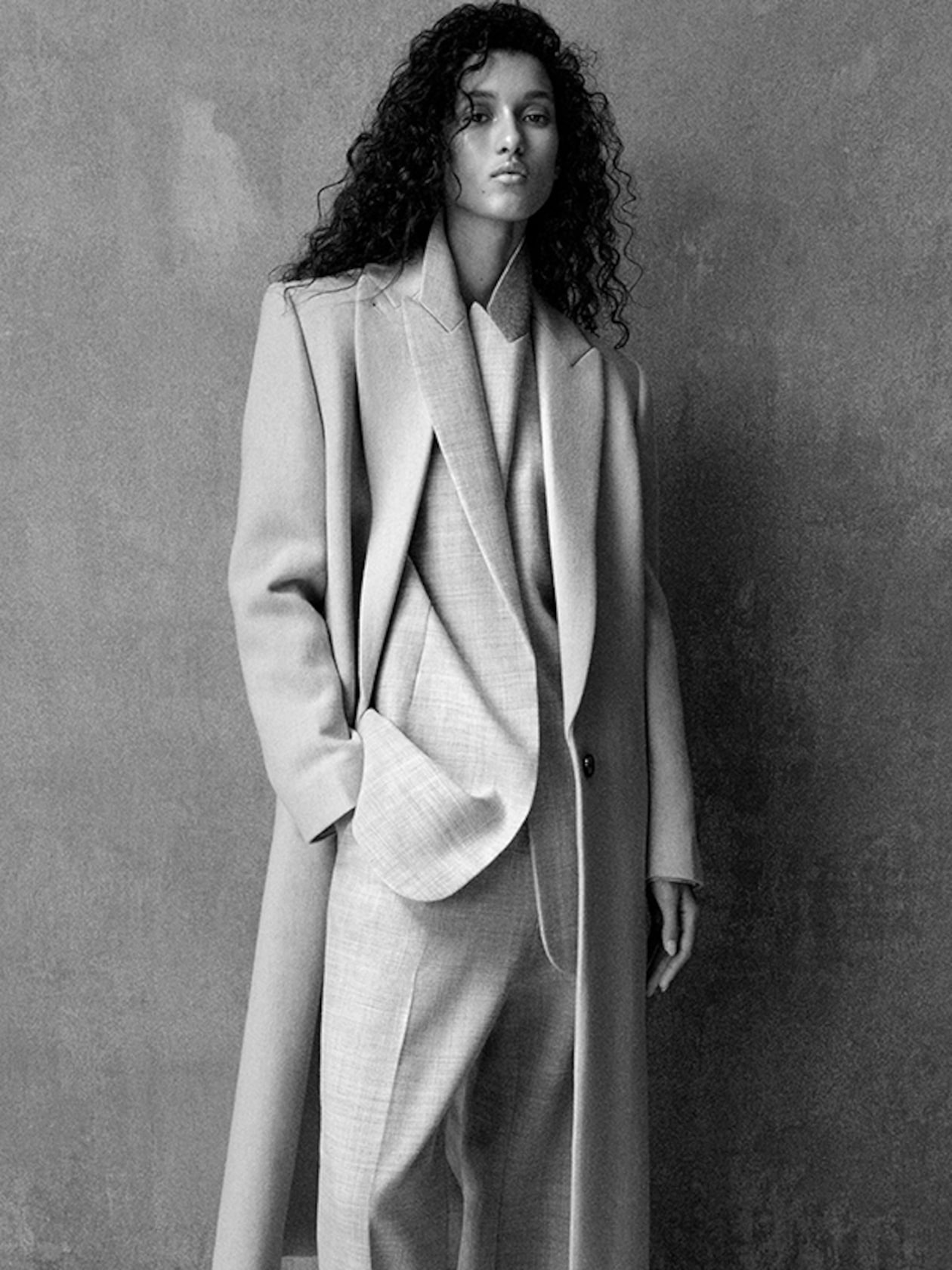 Woman with curly hair in a long coat and suit stands against a textured wall in a black and white photo.