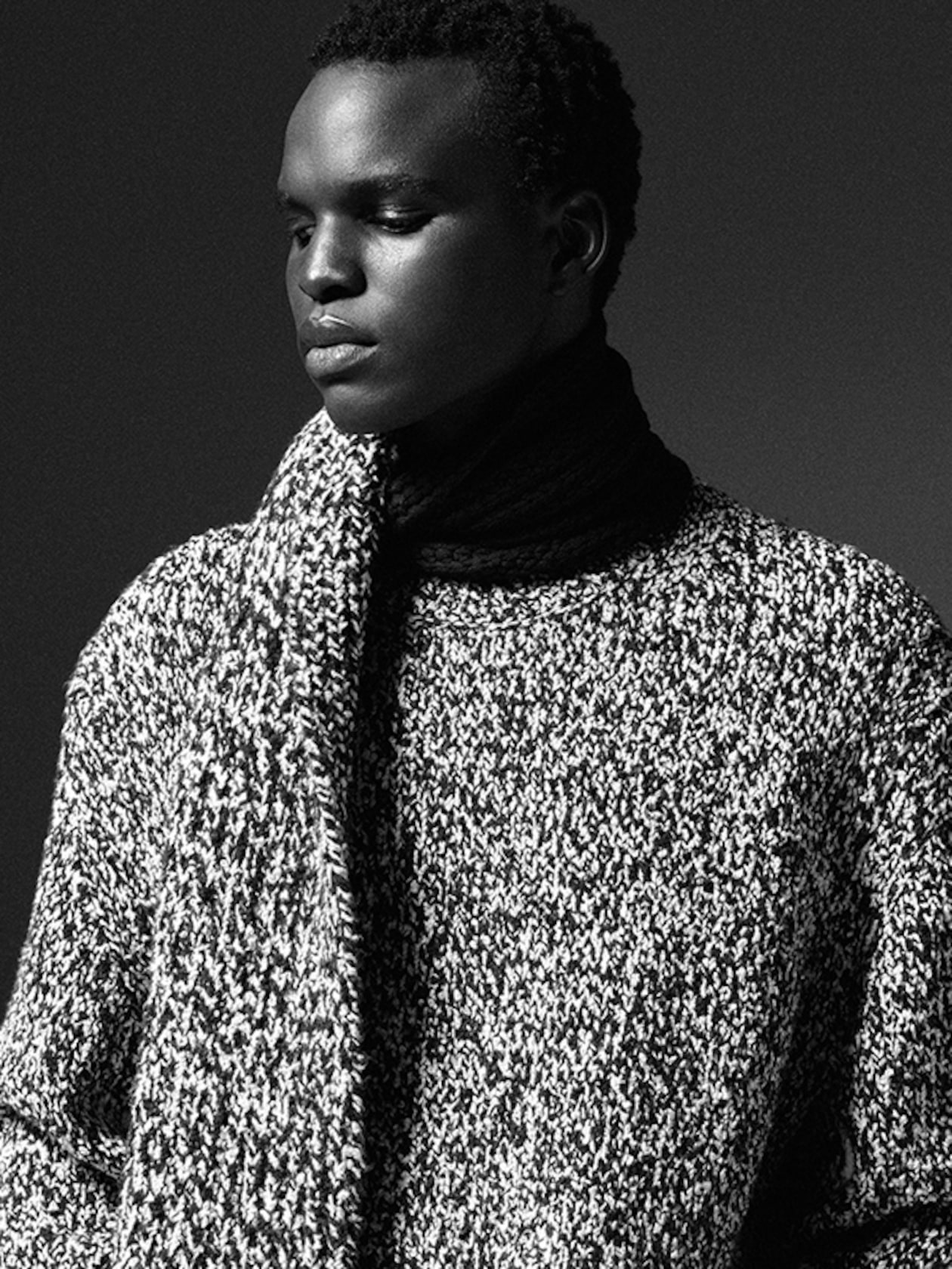 Black and white portrait of a person wearing a textured knit sweater and scarf, looking thoughtfully to the side.