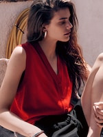 Placeholder for: A lifestyle portrait of a woman seated outdoors, wearing a vibrant red V-neck sleeveless cardigan vest with tonal buttons and black drawstring trousers. She is styled with chunky silver hoop earrings and a matching silver bangle. The sunlit scene features her in profile with long, wavy hair, leaning against a textured white wall with a yellow garden hose visible in the background.