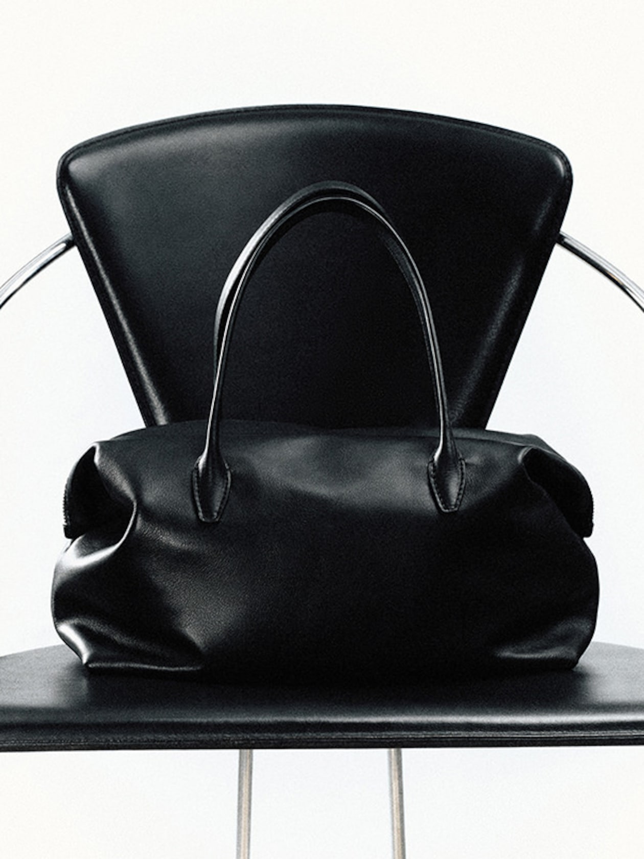 A black leather handbag with handles sits on a matching black chair with a modern design.
