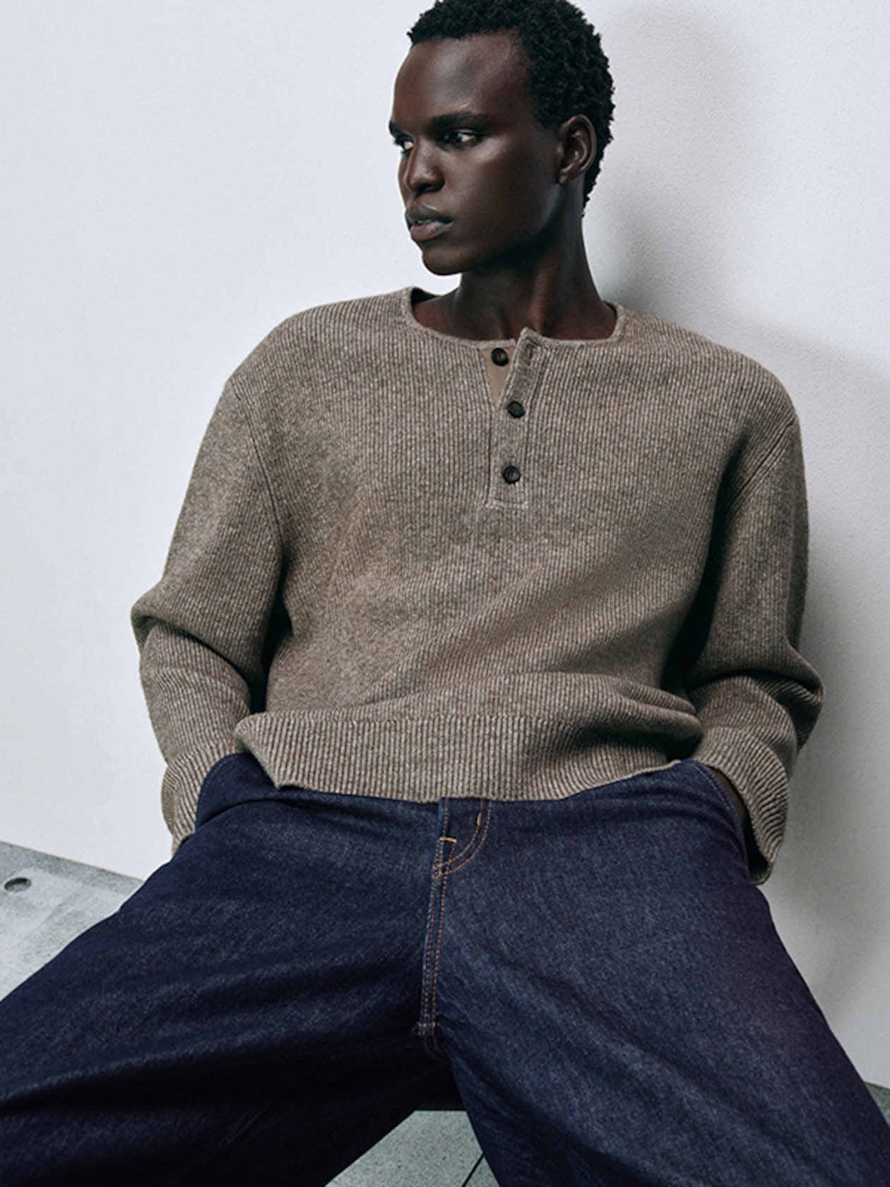 Man in a beige knit sweater and dark jeans leans against a white wall, looking to the side. Casual and relaxed pose.