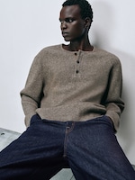 Placeholder for: Man in a beige knit sweater and dark jeans leans against a white wall, looking to the side. Casual and relaxed pose.