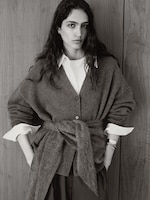 Placeholder for: Woman in a belted cardigan with a white shirt underneath, standing against a wooden background, with hands in pockets. Black and white photo.