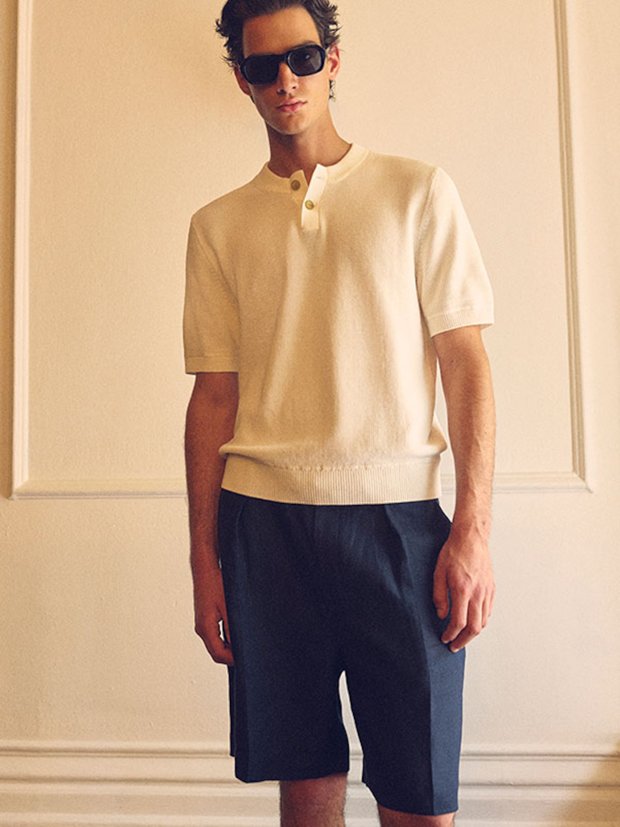 A person wearing sunglasses, a light-colored short-sleeve sweater, and dark shorts stands in a well-lit room with a neutral background.