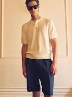 Placeholder for: A person wearing sunglasses, a light-colored short-sleeve sweater, and dark shorts stands in a well-lit room with a neutral background.