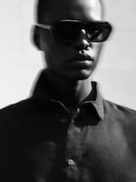 Placeholder for: Person wearing dark sunglasses and a button-up shirt, standing against a blurred, monochrome background.