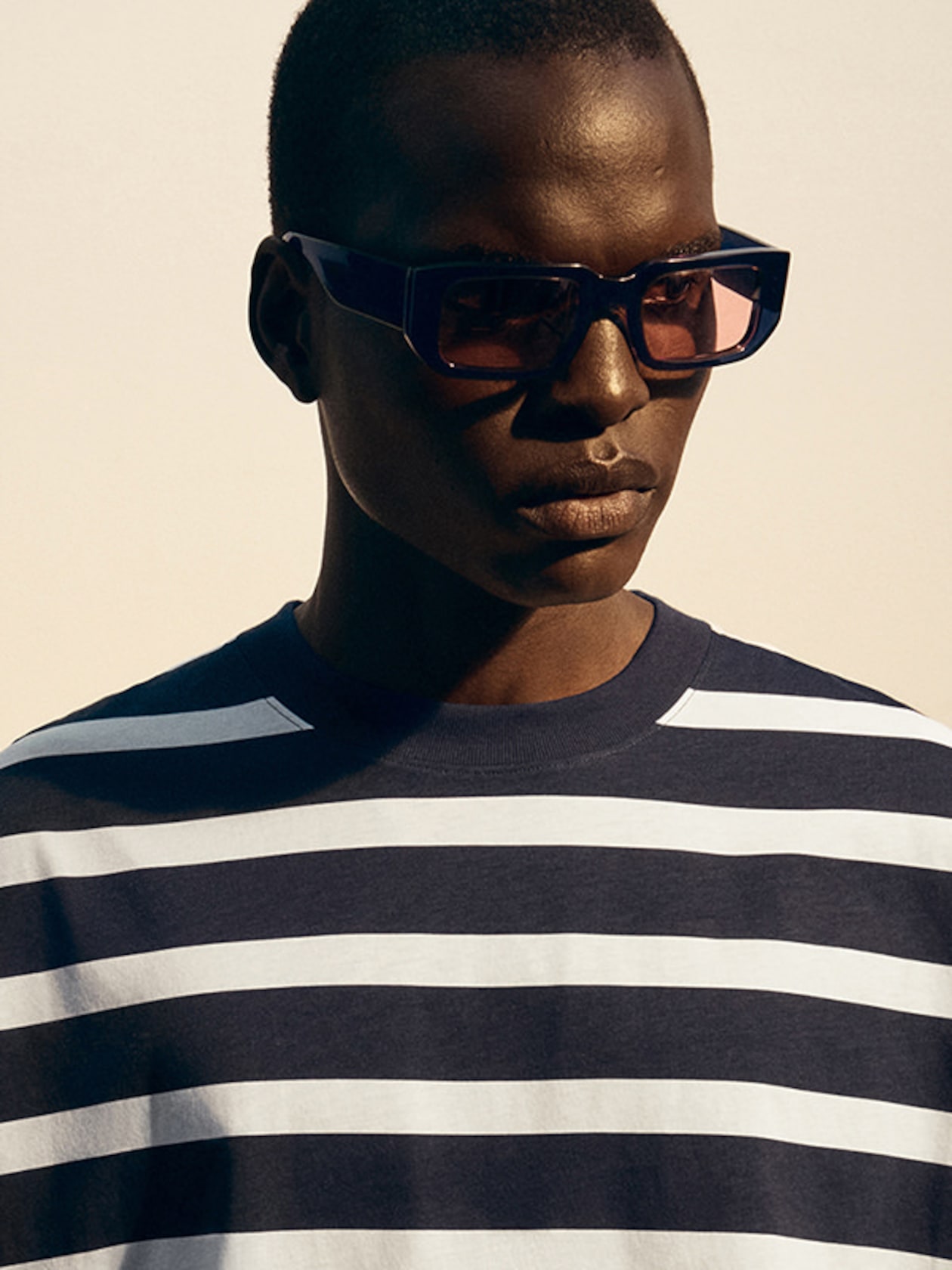 Person wearing rectangular sunglasses and a navy and white striped shirt, looking to the side against a neutral background.