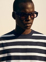 Placeholder for: Person wearing rectangular sunglasses and a navy and white striped shirt, looking to the side against a neutral background.