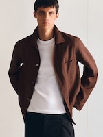 Placeholder for: A person wearing a brown button-up jacket over a white T-shirt and black pants, standing against a plain background with hands in pockets.