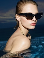 Placeholder for: A person with slicked-back hair wearing cat-eye sunglasses and a gold necklace, partially submerged in a pool against a dusky sky.