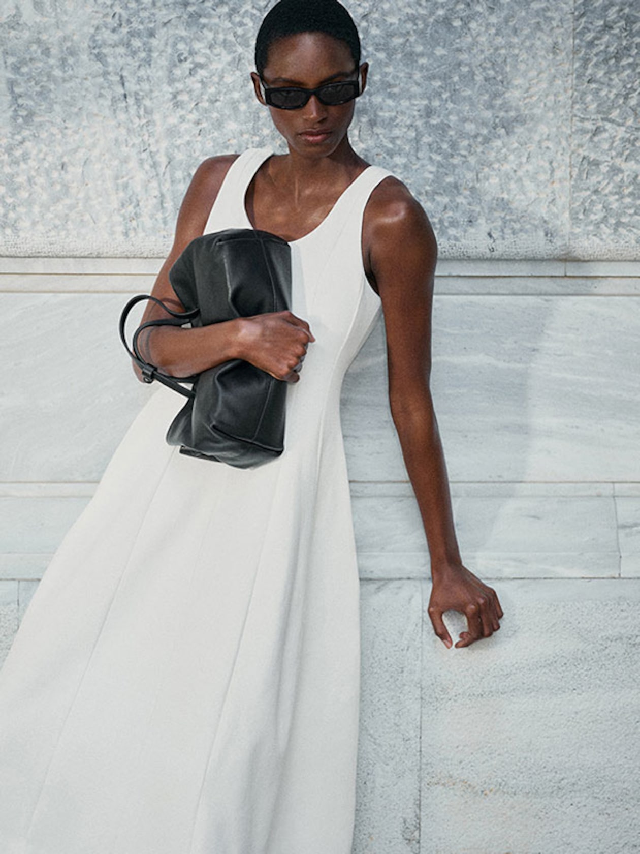 Person wearing a white sleeveless dress and sunglasses, holding a black bag, leaning against a marble wall.
