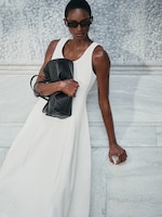 Placeholder for: Person wearing a white sleeveless dress and sunglasses, holding a black bag, leaning against a marble wall.