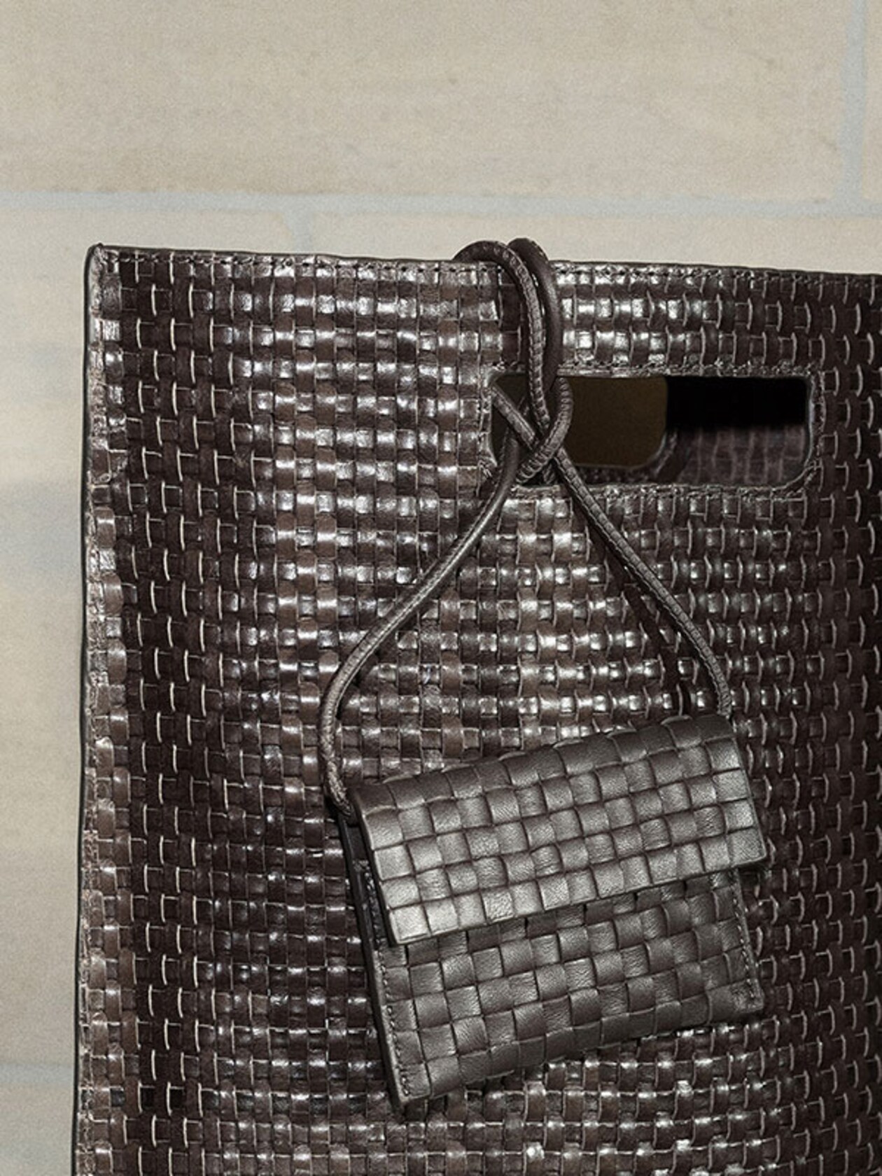 A close-up of a large dark brown woven leather tote bag featuring a rectangular cutout handle. A matching mini woven leather pouch with a thin crossbody strap is draped over the handle. Both bags showcase a glossy, intricate basket-weave texture.