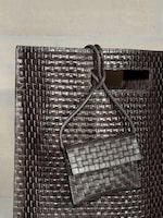 Placeholder for: A close-up of a large dark brown woven leather tote bag featuring a rectangular cutout handle. A matching mini woven leather pouch with a thin crossbody strap is draped over the handle. Both bags showcase a glossy, intricate basket-weave texture.