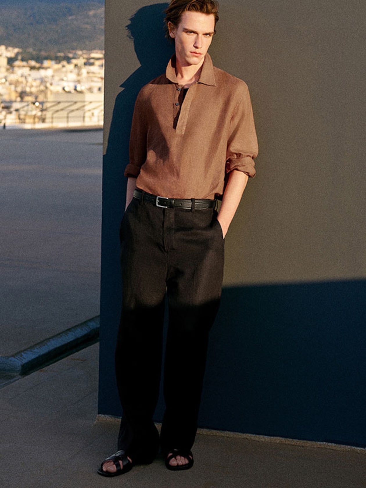 A person in a brown shirt and black pants leans against a gray wall, with hands in pockets and wearing sandals, under a clear sky on a rooftop.