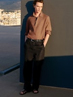 Placeholder for: A person in a brown shirt and black pants leans against a gray wall, with hands in pockets and wearing sandals, under a clear sky on a rooftop.