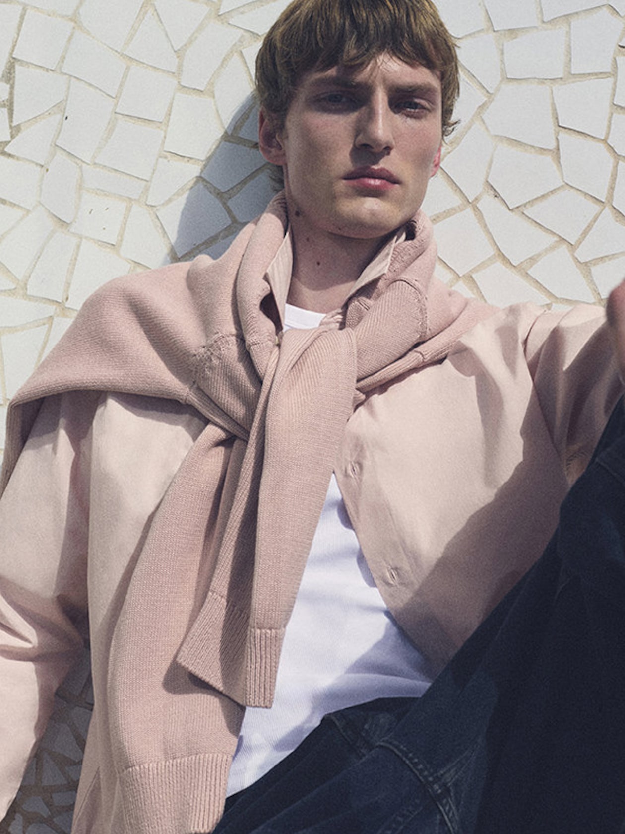Person wearing a pastel pink sweater tied over a white shirt, sitting against a mosaic-patterned wall in sunlight.