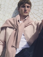 Placeholder for: Person wearing a pastel pink sweater tied over a white shirt, sitting against a mosaic-patterned wall in sunlight.