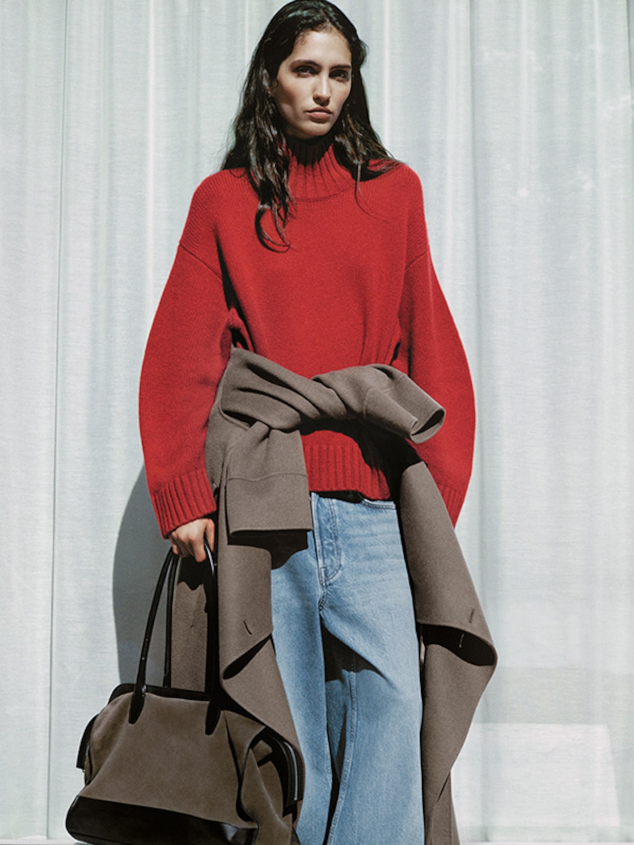 Person in a red sweater and blue jeans, holding a large brown bag, stands against a white curtain background.