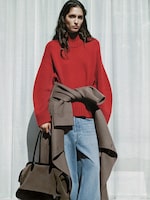 Placeholder for: Person in a red sweater and blue jeans, holding a large brown bag, stands against a white curtain background.