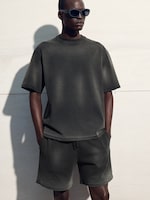 Placeholder for: Person wearing sunglasses, a dark oversized t-shirt, and matching shorts, standing against a light wall with hands in pockets.