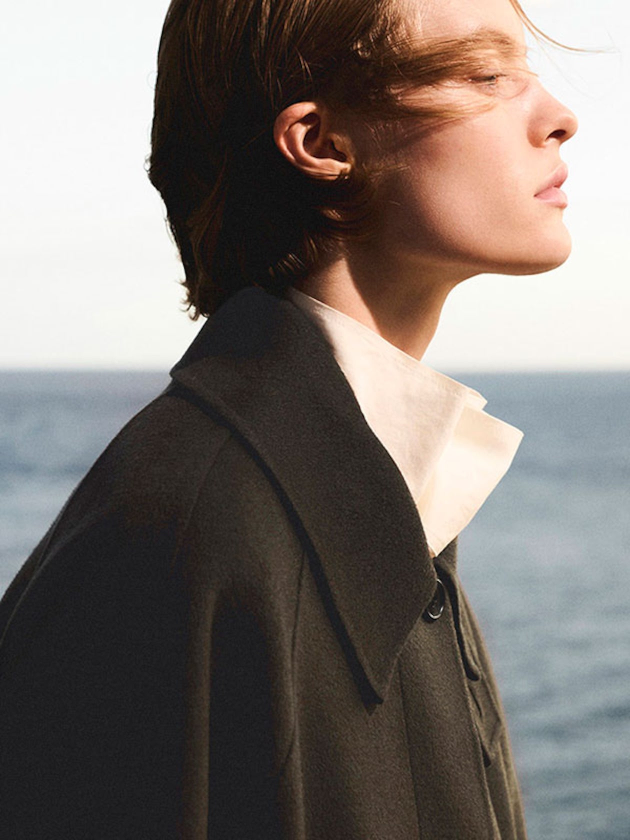 Side profile of a person with short hair, wearing a dark coat and white shirt, standing by the sea with a serene expression.